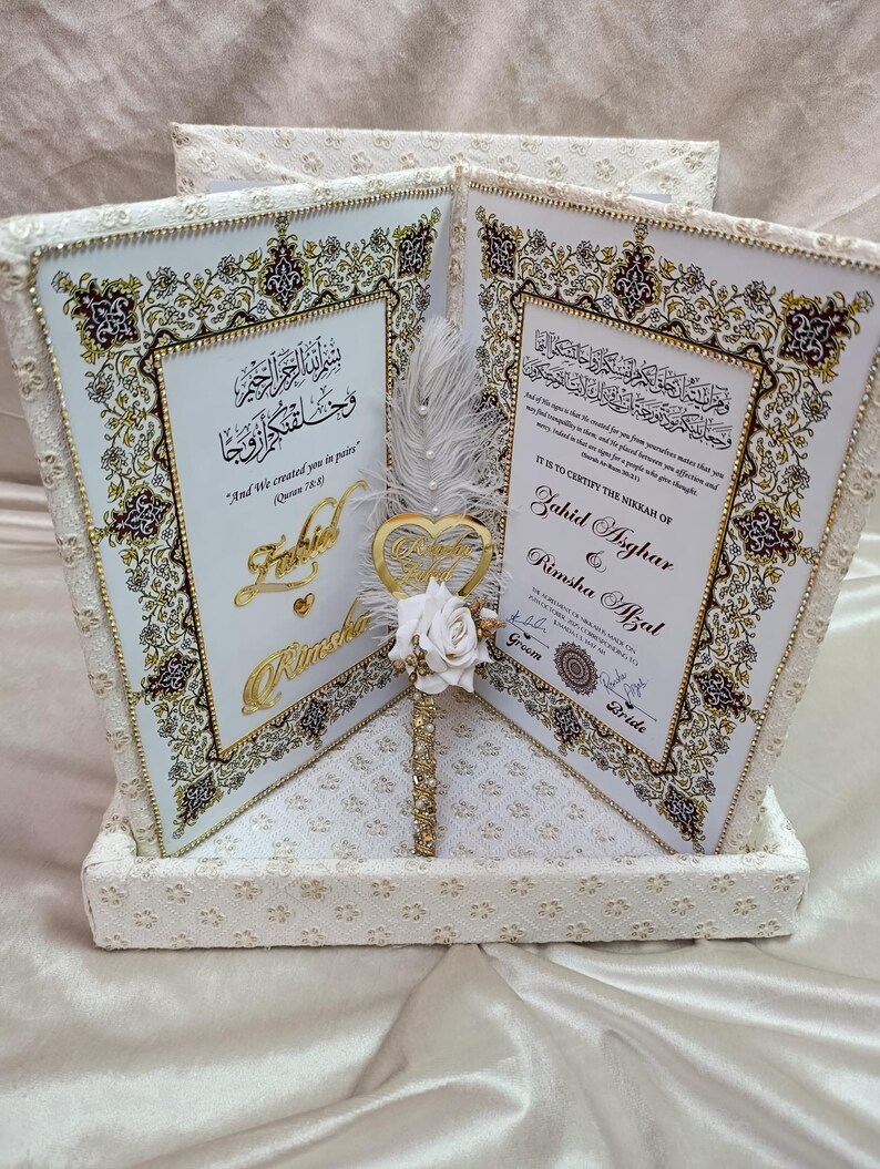 May include: A wedding gift box featuring two framed certificates with ornate gold and brown detailing on a white background. The box itself is white with a lace pattern. A gold heart ornament and white rose are in the center.