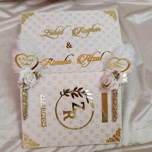 May include: A white decorative box with gold detailing, inscribed with the names "Fahid Asghar" and "Rimsha Afzal" in gold script. The box is decorated with floral and feather accents, and the date "25/10/2025" is visible.