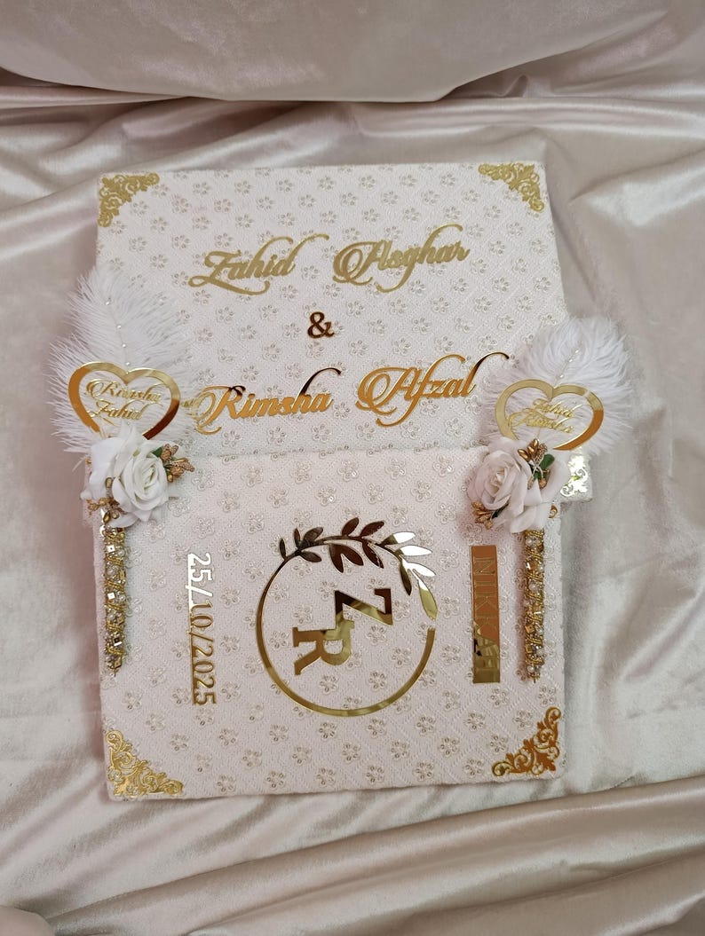 May include: A white decorative box with gold detailing, inscribed with the names "Fahid Asghar" and "Rimsha Afzal" in gold script. The box is decorated with floral and feather accents, and the date "25/10/2025" is visible.
