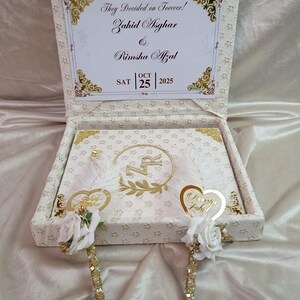 May include: A cream-coloured wedding box with gold detailing and a floral pattern. The box is open, revealing a card with the text "They Decided on Forever!" and the names Zahid Asghar and Rimsha Afzal, with the date 25 October 2025.