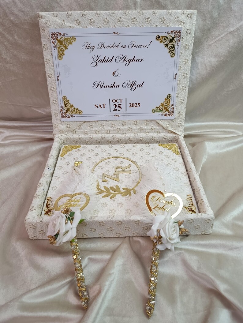 May include: A cream-coloured wedding box with gold detailing and a floral pattern. The box is open, revealing a card with the text "They Decided on Forever!" and the names Zahid Asghar and Rimsha Afzal, with the date 25 October 2025.