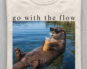 May include: An ivory t-shirt featuring a graphic of an otter wearing black sunglasses and holding an iced coffee while floating in water. The text "go with the flow" is printed in black above the image. The shirt has a crew neckline.