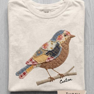 May include: Ivory-colored sweatshirt with a detailed embroidered bird design. The bird features patchwork-style wings in various colors and patterns, perched on a branch. The word "Custom" is written below the branch. The word "Ivory" is written on a label.
