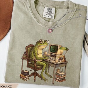 May include: A khaki crew neck t-shirt featuring a graphic of a green frog wearing glasses, sitting in a chair at a desk with a computer, a lamp, and stacks of books. The shirt has a Comfort Colors brand label at the neck and is size S.