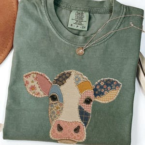 May include: Moss green t-shirt with a patchwork cow face design. The cow's face features various fabric patterns in shades of pink, blue, yellow, and floral prints. A gold necklace with a small pendant rests on the shirt. A brown hat is visible in the upper left corner.