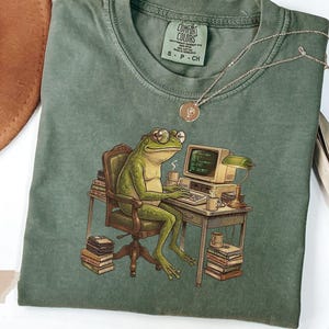May include: Moss green t-shirt with a graphic of a frog wearing glasses and typing on a computer. The frog is seated at a desk with books and a lamp. A gold necklace is draped on the shirt.
