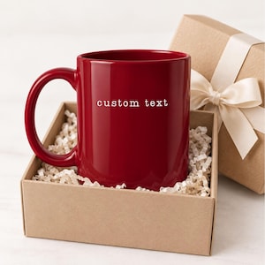 May include: A deep red ceramic mug featuring the white text custom text printed on the front. The mug is nestled inside a square cardboard gift box filled with light-colored paper filler, accompanied by a matching lid with a white ribbon.