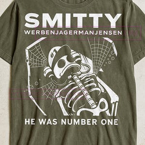 Könnte beinhalten: Olivgrünes T-Shirt mit weißem Grafiktext und Illustration. Der Text oben lautet "SMITTY WERBENJAGERMANJENSEN". Die Illustration zeigt ein Skelett mit Hut und Klarinette. Der Text unten lautet "HE WAS NUMBER ONE".