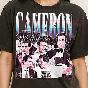 May include: Dark gray t-shirt featuring a graphic of Cameron Winklevoss. The design includes multiple images of Winklevoss, with the name "CAMERON WINKLEVOSS" in a stylized font. The shirt has a vintage, distressed look.