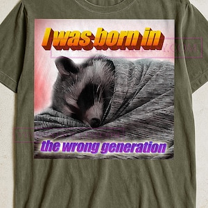 May include: An olive green t-shirt with a graphic of a raccoon wrapped in a blanket. The graphic includes the text "I was born in the wrong generation" in yellow and purple. The raccoon is black, white, and grey.