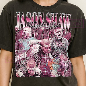 May include: Dark gray t-shirt featuring a graphic of a man with the text "JASON SHAW" in a pink and white retro design. The graphic includes multiple images of the man in various poses, with a vintage aesthetic.