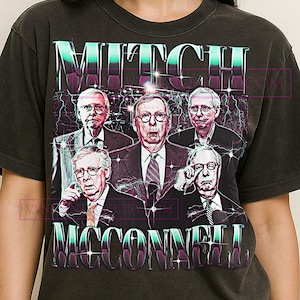Op de afbeelding: Donkergrijs t-shirt met een retro graphic van Mitch McConnell en de tekst "MITCH MCCONNELL". Het ontwerp toont een portret van Mitch McConnell en andere mannen in pakken. De tekst is in een groen-paarse gradiënt.