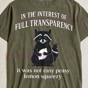 Puede incluir: Camiseta verde oliva con un dibujo de un mapache sosteniendo un limón. El texto en la camiseta dice "IN THE INTEREST OF FULL TRANSPARENCY" encima del mapache y "it was not easy peasy lemon squeezy" debajo.