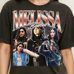 May include: Dark gray t-shirt featuring a graphic of the name "MELISSA" in a vintage style, with the last name "Barrera" below. The graphic includes multiple images of a woman, with lightning bolt accents.