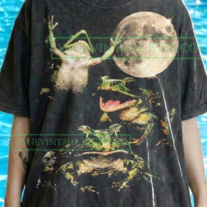 Può includere: T-shirt grigio scuro con una grafica di tre rane e una luna piena. Le rane sono verdi e bianche, con una rana che alza le braccia. Il testo "Original design by VYNE.VINTAGE" è stampato in basso.