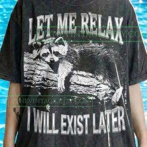 May include: Dark gray t-shirt featuring a black and white raccoon illustration resting on a log. The text "LET ME RELAX I WILL EXIST LATER" is printed above and below the raccoon. The shirt has a vintage style.