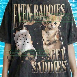 Puede incluir: Camiseta gris oscuro con el texto "EVEN BADDIES GET SADDIES" y un gráfico de tres gatitos y un zapato blanco. El diseño incluye un gato negro y dos gatitos atigrados naranjas.