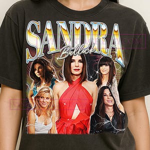 May include: Dark gray t-shirt featuring a graphic of Sandra Bullock with the text "SANDRA BULLOCK" in a vintage style. The graphic includes multiple images of the actress in various poses and outfits.