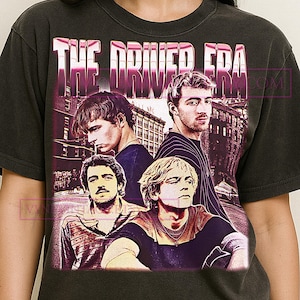 May include: Dark gray t-shirt featuring a graphic of four men with the text "THE DRIVER ERA" in a pink and white font. The image has a vintage, faded look, with a city backdrop.