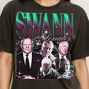 Può includere: T-shirt grigio scuro con un design grafico. Il design presenta il nome "SWANN" in grandi lettere stilizzate, con diversi ritratti di uomini sotto e la parola "Senator". Il design ha un'estetica vintage.