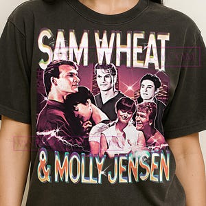 May include: Dark gray t-shirt featuring a graphic of several people with the text "SAM WHEAT & MOLLY JENSEN" in a vintage style. The graphic has a purple and pink color scheme.