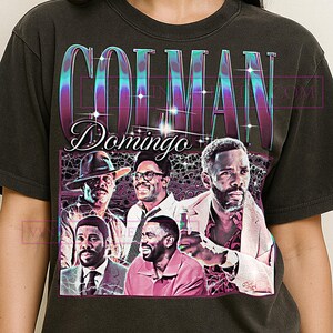 May include: Dark gray t-shirt featuring a vintage-style graphic of Colman Domingo and other men. The design includes the name "COLMAN DOMINGO" in a retro font with a multi-colored gradient. The graphic showcases several images of the actor.