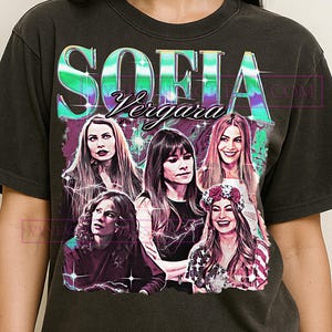 Puede incluir: Camiseta gris oscuro con un gráfico de Sofía Vergara y el texto "SOFIA Vergara" en un diseño vibrante y multicolor. El gráfico incluye imágenes de varias mujeres. La camiseta es de cuello redondo.