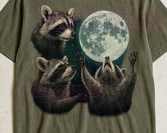 May include: Olive green t-shirt featuring a graphic of three raccoons gazing at a full moon. The raccoons are in shades of gray and black, with one raising its paws towards the moon. The moon is a bright white color.
