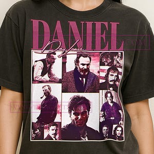 Può includere: T-shirt grigio scuro con una grafica rosa che mostra il nome "DANIEL Day-Lewis" e una griglia di immagini. Le immagini sono ritratti dell'attore in vari ruoli.