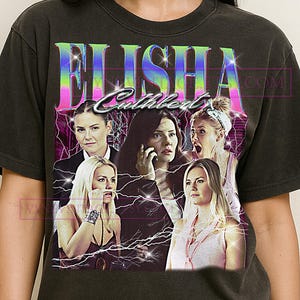 May include: Dark gray t-shirt featuring a graphic of Elisha Cuthbert with a retro design. The design includes the name "Elisha Cuthbert" in a colorful, stylized font, along with multiple images of the actress. The shirt has a vintage, distressed look.