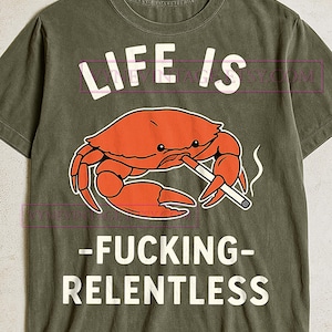 Puede incluir: Camiseta verde oliva con un gráfico de un cangrejo naranja fumando un cigarrillo. El texto "LIFE IS" está encima del cangrejo, y "-FUCKING- RELENTLESS" debajo, todo en blanco.