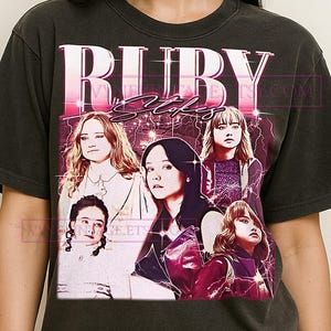 Puede incluir: Camiseta gris oscuro con un diseño gráfico retro. El diseño presenta el texto "RUBY Stokes" en rosa y blanco, junto con un collage de cinco mujeres y un efecto de rayo rosa.
