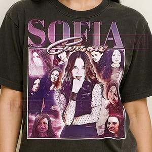 May include: Dark gray t-shirt featuring a graphic of Sofia Carson with the name "SOFIA Carson" in a vintage style. The design includes multiple images of Sofia Carson. The shirt is a crew neck.