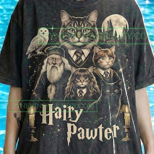Puede incluir: Camiseta gris oscuro con un diseño caprichoso con personajes de gatos vestidos con uniformes escolares, un búho y el texto "Hairy Pawter". El diseño incluye una luna llena, un castillo y velas. La camiseta es de diseño vintage.