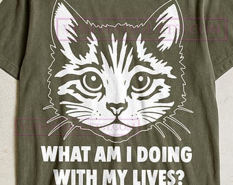 Könnte beinhalten: Olivgrünes T-Shirt mit einem weißen Katzenkopf-Motiv. Darunter steht in weißen Großbuchstaben der Text "WHAT AM I DOING WITH MY LIVES?". Das T-Shirt hat einen Rundhalsausschnitt und kurze Ärmel.
