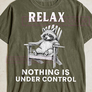May include: Olive green t-shirt featuring a relaxed raccoon in a wooden chair. The text "RELAX" is above the raccoon, and "NOTHING IS UNDER CONTROL" is below. The design is in white.
