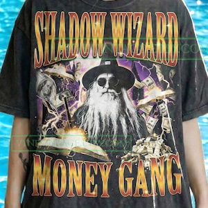 May include: Dark gray t-shirt with the text "SHADOW WIZARD MONEY GANG" in gold and red lettering. The graphic features a wizard with sunglasses, a long white beard, and money-themed imagery. The design is by VYNE.VINTAGE.