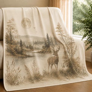 May include: A cream-coloured throw blanket featuring a sepia-toned landscape illustration. The scene depicts a stag standing near a riverbank, surrounded by pine trees, tall grasses, and cattails, with a full moon visible in the sky above.