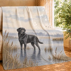 Peut inclure: Un plaid doux présentant une illustration imprimée d'un chien Labrador noir debout dans une eau peu profonde. La scène comprend de hautes herbes dorées au premier plan, un lac calme et un ciel doux avec trois oiseaux volant au loin.
