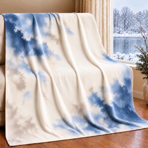 May include: A soft throw blanket featuring a cream-colored base with a cloud-like pattern in shades of blue and grey. The design creates a gradient effect across the fabric, which is draped over furniture, showcasing a smooth, plush texture.
