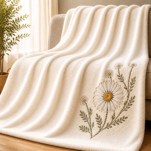 May include: Cream-colored, plush throw blanket with an embroidered daisy design. The blanket is draped over a light-colored sofa. The daisy design features white petals, a yellow center, and green stems and leaves.