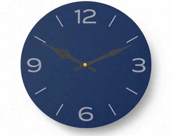 May include: A round, navy blue wall clock with white numerals at 3, 6, 9, and 12 o'clock. The clock hands are black, and the center is gold. The clock is a simple, modern design.