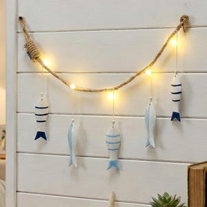 May include: A nautical-themed string of lights with small, warm white bulbs. The lights are strung along a thick, natural rope, and hang from a white wooden wall. Five wooden fish ornaments, painted white with blue stripes and fins, are suspended from the rope.