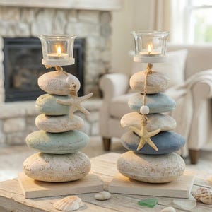 Peut inclure: Deux bougeoirs décoratifs. Chacun présente une pile de pierres peintes dans des tons beiges, bleus et verts, surmontée d'un photophore en verre. Des étoiles de mer et des coquillages ajoutent un thème côtier.