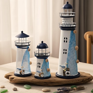 Puede incluir: Tres figuras decorativas de faros de diferentes tamaños. Los faros son blancos con detalles azules, incluyendo un diseño de red azul y tapas azul oscuro. Cada faro presenta pequeñas ventanas y elementos decorativos como gaviotas y conchas.