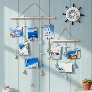 Puede incluir: Expositor de fotos de madera con impresiones de arquitectura griega y paisajes marinos. Decorado con conchas marinas, anclas y estrellas azules. Una decoración de timón náutico adorna la pared. El conjunto está fijado a una pared azul.
