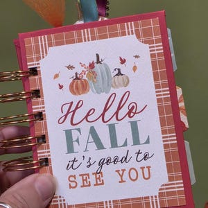 Könnte beinhalten: Ein kleines Notizbuch mit Spiralbindung, rotem Einband mit Karomuster und einem zentralen Etikett. Das Etikett zeigt Illustrationen von drei Kürbissen und den Text "Hello FALL it's good to SEE YOU" in roter, grüner und oranger Schrift.
