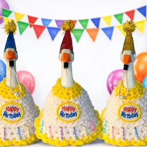 May include: Three goose-shaped bottle stoppers wearing birthday hats and crocheted cake-shaped outfits. Each outfit has a "Happy Birthday" patch and candle accents. Colorful balloons and a banner decorate the background.
