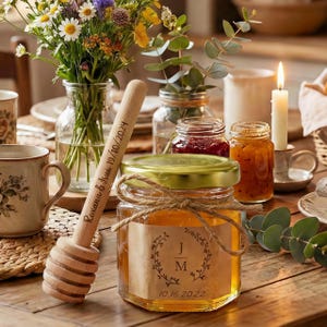 Op de afbeelding: Een pot honing met een gouden deksel en een etiket met de initialen "J M" en de datum "10.16.2022". Een houten honingdipper en potten jam zijn ook zichtbaar. Een boeket bloemen en een brandende kaars staan op de achtergrond.