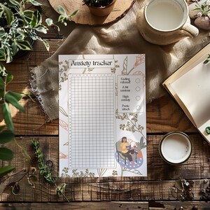 May include: Anxiety tracker journal with a grid for tracking feelings. The journal has floral and botanical illustrations in green and peach. The words "Anxiety tracker" are at the top. A person is illustrated reading a book in a chair.